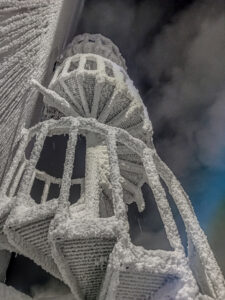 The image shows a spiral staircase, completely covered in frost or snow, likely in a freezing outdoor environment. The thick coating of ice gives the structure a surreal, almost otherworldly appearance. The staircase&#039;s metal framework is barely visible beneath the heavy layer of frost, which seems to have accumulated in a uniform, crystalline fashion, forming textured surfaces. The dark, cloudy sky in the background adds to the cold and harsh atmosphere, making the image feel like it was taken in extreme winter conditions. The perspective looks up from the base of the staircase, emphasizing its height and the intricate, frozen details.