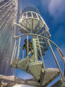 Long and icy circular stairs leading up the exterior of an icy industrial building. A man with a shovel and high visibility clothing is standing a few steps up the stairs looking back at the viewer.