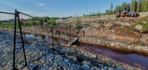 The image shows a construction site, with a fenced walkway leading down to a rocky ditch. The ditch is filled with water that appears to be brown or murky. The area surrounding the ditch is made up of exposed dirt, rocks and gravel. There are three sections of pipeline in the background of the image, on the bank of the ditch, as well as numerous trees in the background. The scene appears to be a work in progress, and the ditch may be part of a drainage system for the site.