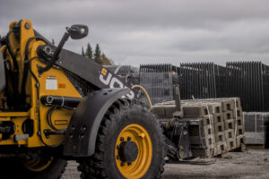 The image shows a close-up of a yellow and black wheel loader, in an outdoor industrial setting. The vehicle is transporting a stack of concrete blocks, which are bound together with plastic straps. The tires of the vehicle are large, rugged, and covered in dirt, indicating that it&#039;s suited for heavy-duty work, likely in construction or logistics.  In the background, there are neatly stacked metal or concrete slabs, and the setting appears overcast with a cloudy sky, giving the scene a cold, industrial atmosphere. Trees can be seen in the distance, suggesting that this is taking place near a construction site or storage yard in a semi-rural area.