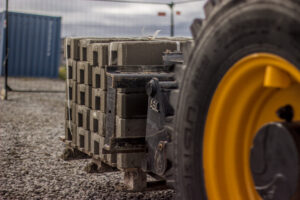 A wheel loader is transporting a pallet of concrete feet closer to where there is some black modular fence getting built.