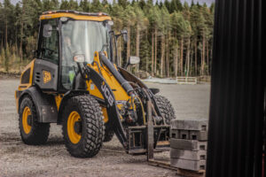 The image features a JCB wheel loader positioned in an outdoor setting, surrounded by a gravel surface and a backdrop of trees. The machine is placing the forks under a pallet of concrete blocks. The overall atmosphere is that of a work site.