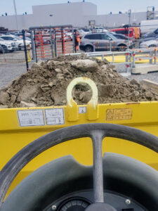 The image shows a close-up of a yellow construction vehicle&#039;s dashboard and a large pile of dirt in front of it. In the background, there are parked cars and a construction site with various structures and activity. The focus is on the vehicle&#039;s controls and the loader&#039;s hook, which is highlighted by the dirt pile.